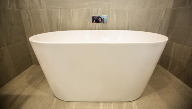 A freestanding white oval bathtub positioned against a wall finished in large-format taupe-grey stone-look tiles, with matching tiled flooring and a small wall-mounted control or fixture above the bath. The bathroom has a clean, contemporary and understated appearance.