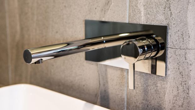Close-up of a wall-mounted chrome bath mixer tap with a cylindrical spout and lever handle, set against a grey textured tiled wall with a dark rectangular backing plate. The curved edge of a white bathtub is visible in the foreground.