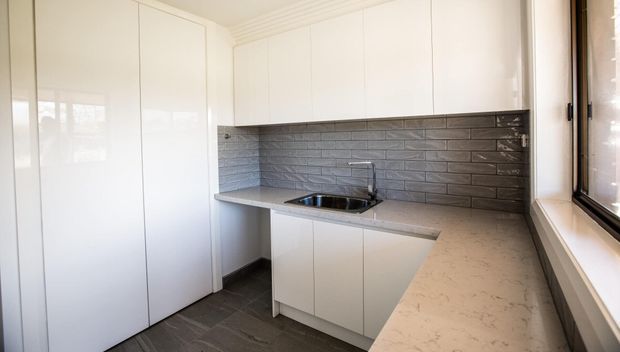 Compact modern kitchen with white full-height cabinetry, a grey textured subway-tile splashback, dark grey tiled flooring, and pale stone-look benchtops. The layout includes an L-shaped work surface, integrated sink, and a large window providing natural light.