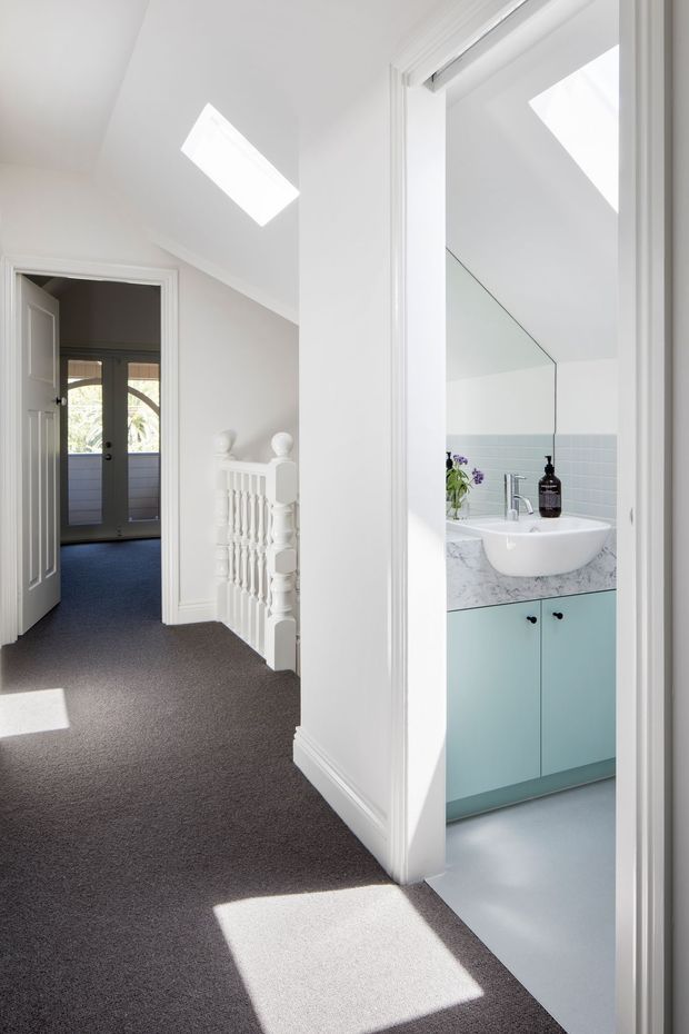 Bright white-painted upstairs hallway or landing with dark grey carpet, traditional balustrade and trim, skylights, and an open doorway revealing a compact bathroom with pale blue tiled walls, a marble-look vanity benchtop, white basin, and pale aqua cabinetry. A glazed exterior door is visible at the end of the hall.