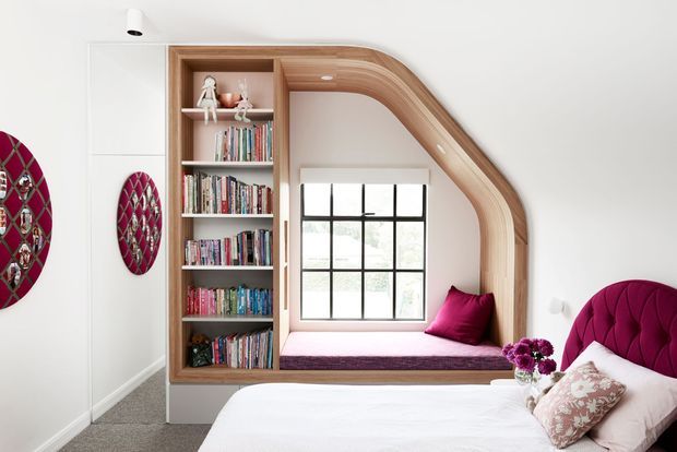 Bright, contemporary bedroom featuring a built-in timber-framed window seat and reading nook integrated beside a tall open bookshelf. The room has a plush deep magenta upholstered bedhead, matching cushion, patterned throw pillows, white bedding, neutral carpet, and decorative magenta wall mirrors. A black-framed gridded window provides natural light.