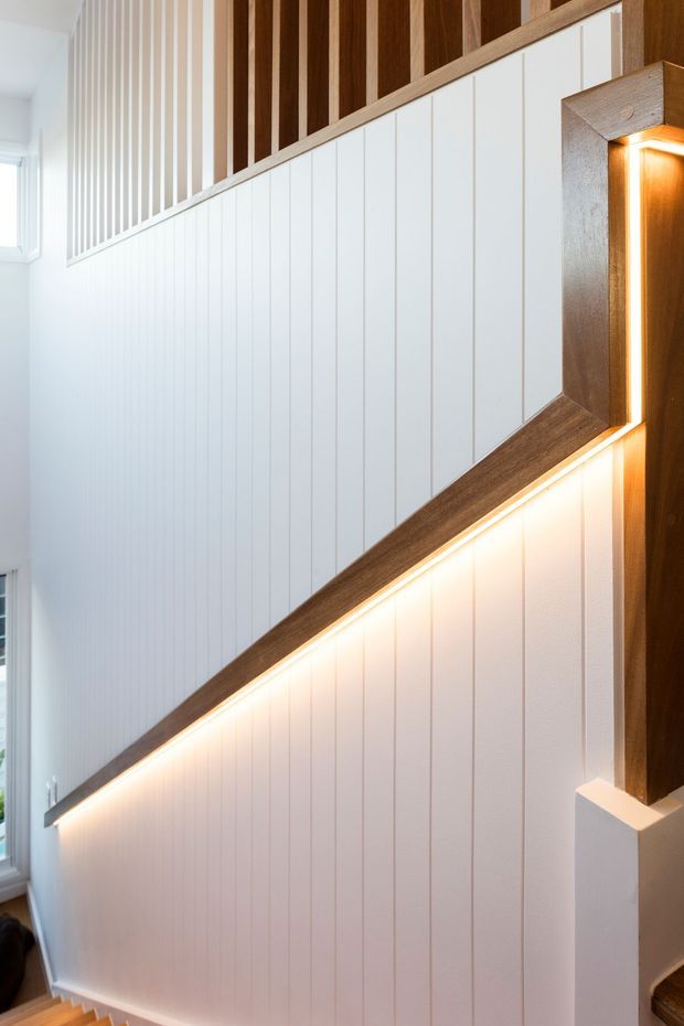 Close-up interior view of a stairway with white vertical panelled walls, warm timber trim and a timber slatted balustrade. Integrated warm LED strip lighting runs beneath the diagonal handrail or wall trim, creating a refined architectural feature. A tall window provides natural light at the side.