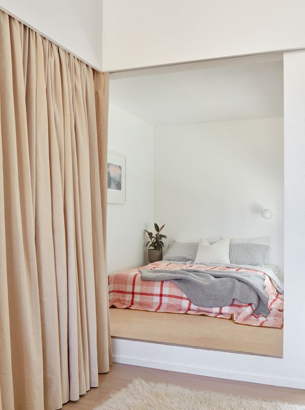 A bright, minimalist bedroom viewed through a wide opening, with a low built-in platform bed dressed in layered white, grey, and red plaid bedding. Full-height beige curtains define the left side, while white walls, pale timber flooring and platform surfaces, a small plant, framed artwork, and a wall sconce create a soft, calm interior.