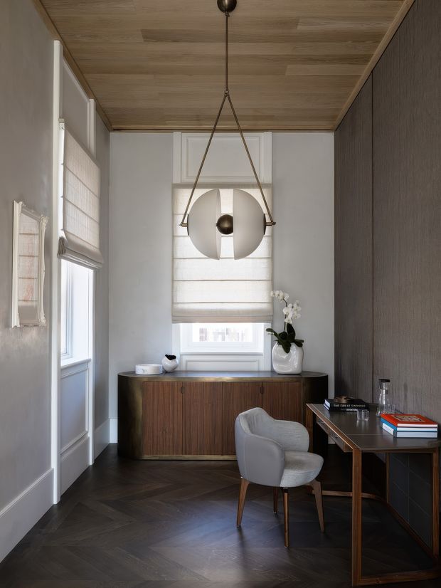 A refined, compact home office with a dark herringbone timber floor, pale paneled walls, a richly grained timber sideboard, and a slim timber writing desk. A grey upholstered chair sits at the desk, while a sculptural pendant hangs beneath a warm timber-lined ceiling. Tall windows with woven Roman blinds provide soft natural light, and a dark textured feature wall adds depth.