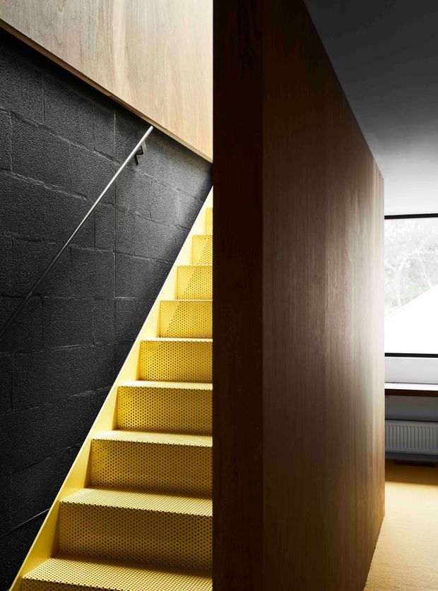 Interior architectural photograph focused on a steep, compact stairway with vivid yellow perforated metal treads and risers. The stair is enclosed by a dark painted concrete-block wall on one side and a tall vertical timber-clad partition on the other. A slim metal handrail follows the wall, while a bright window and part of an adjoining room are visible beyond the partition. The composition is graphic and contemporary, with strong contrast between yellow, black, and warm wood.