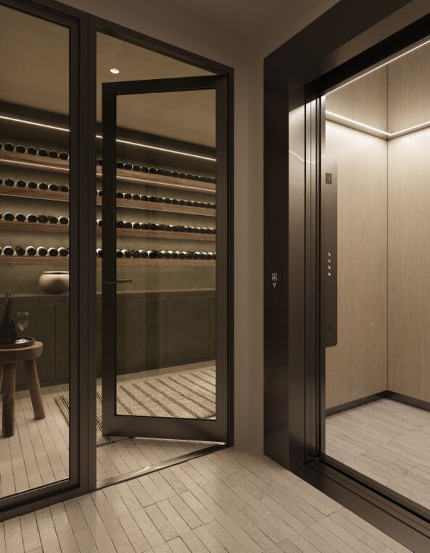 A refined, dimly lit interior showing a compact wine room behind black-framed glass doors, with multiple illuminated wine-bottle shelves and dark built-in cabinetry. Beside it is a modern elevator with warm wood-look wall panels, integrated lighting, and a dark metal surround. Pale timber-look flooring runs through the space, creating a luxurious, contemporary atmosphere.
