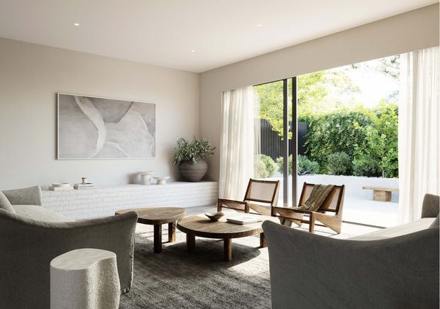 A refined, light-filled living room with neutral beige walls, a white painted brick fireplace or low built-in feature, sculptural grey lounge chairs, a round timber coffee table, and woven timber-framed armchairs. Full-height glazing with sheer curtains opens to a planted outdoor area. The room has a restrained contemporary aesthetic with natural textures and soft, warm neutral tones.