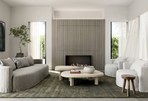 A refined contemporary living room with a large curved sectional sofa, sculptural coffee tables, soft white furnishings, and a linear fireplace set into a full-height vertically ribbed grey feature wall. Sheer curtains frame tall windows, while a muted textured rug and potted indoor tree add warmth and softness.