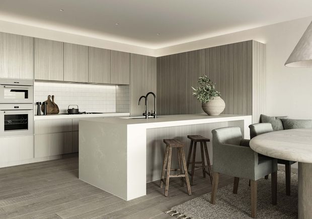 Contemporary open-plan kitchen and dining area with full-height taupe wood-grain cabinetry, integrated appliances, a pale stone island with a waterfall edge, subtle white tiled splashback, and dark timber stools. A round dining table with upholstered chairs sits beside the island, creating a refined, neutral-toned interior.