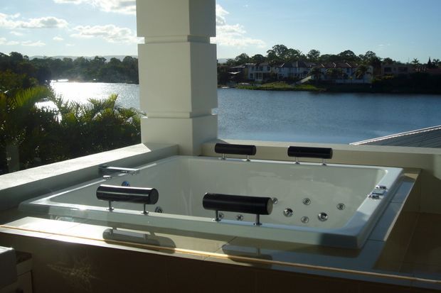 A large white spa bath with multiple jets is installed on a covered balcony or terrace overlooking a calm lake or waterfront. White structural columns, tiled surfaces and dark metal spa fittings frame the outdoor setting, with houses and trees visible across the water.