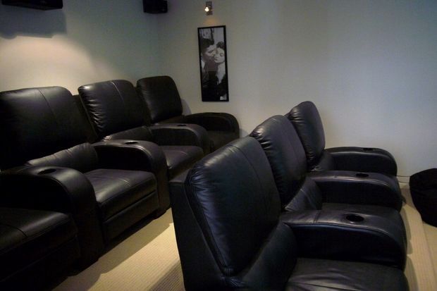 A compact private home cinema with rows of black leather reclining armchairs, integrated cup holders, dim wall lighting, and a framed monochrome artwork. The room has pale walls and a light carpeted floor, creating a dark, intimate viewing atmosphere.