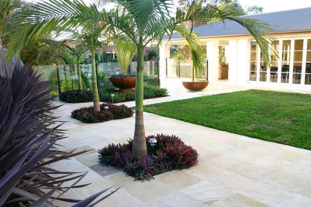 A bright landscaped courtyard beside a single-storey building, featuring pale stone paving, manicured lawn, tropical palms, sculptural planters, low ornamental plantings, and extensive white-framed glazing beneath a pitched metal roof.