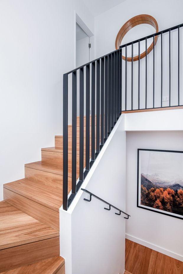 Bright contemporary stairway with warm timber treads and flooring, white walls, and slim black metal balusters and handrails. The stair rises to an upper landing beside a white door, with a large circular timber wall feature and framed landscape artwork.