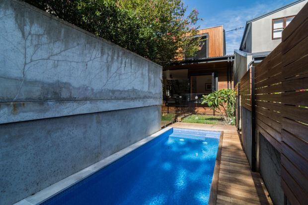 A narrow urban courtyard with a compact rectangular swimming pool bordered by timber decking. Concrete retaining walls and a horizontal timber privacy fence frame the space, leading toward a contemporary multi-level house with timber-clad upper sections, dark-framed glazing, and an outdoor dining or living area. Bright sunlight creates a fresh, modern atmosphere.