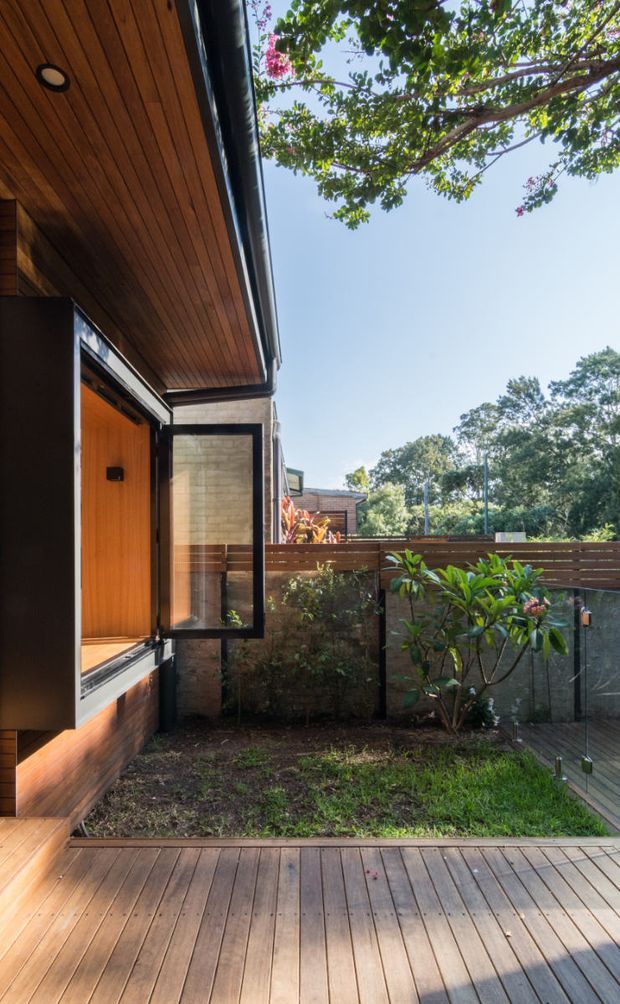 A residential exterior courtyard or deck viewed from a timber-clad building. The scene features a warm timber soffit and wall, black-framed open glazing, timber decking, a horizontal timber privacy fence, concrete masonry planter walls, and landscaped planting beneath mature trees. The image has a bright, leafy, contemporary outdoor atmosphere.