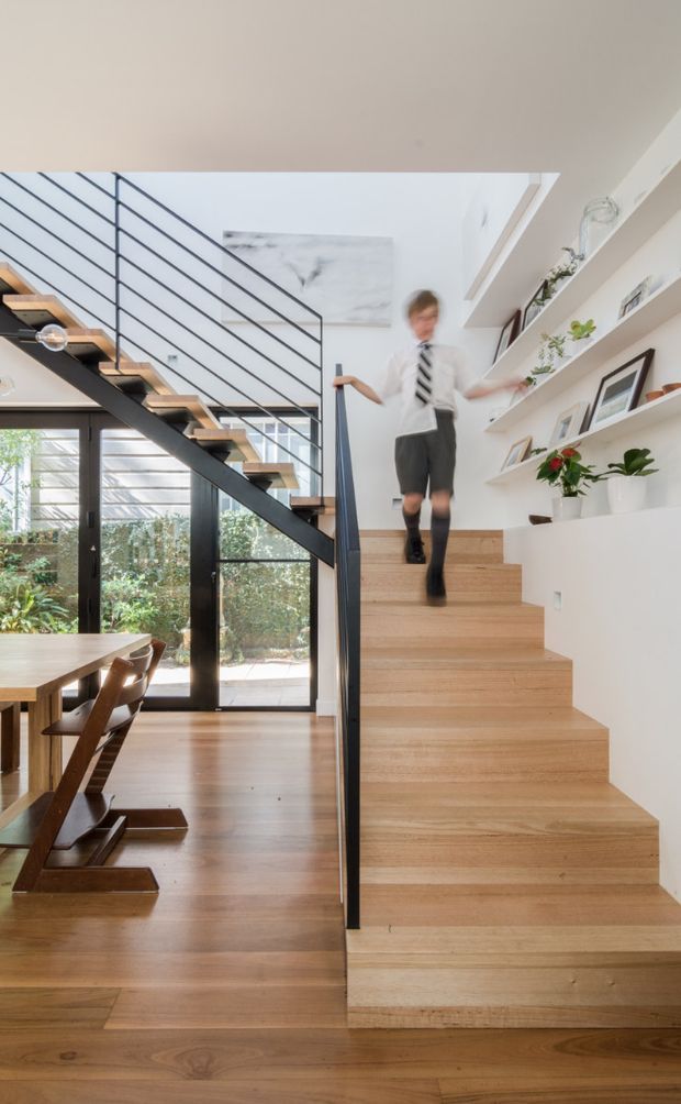 Bright contemporary residential interior showing a timber staircase with broad integrated timber treads and a slim black metal balustrade. The staircase rises beside a dining area with a timber table and chairs, full-height glazed doors, and abundant natural light. White walls and built-in display shelving hold plants, framed artwork, and decorative objects. A child is blurred while descending the stairs.