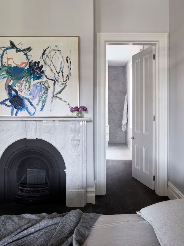 Elegant bedroom interior viewed toward an open bathroom doorway. The foreground features a neatly made bed with white bedding and a grey throw, while the room is anchored by a classic white marble fireplace with a dark cast-iron insert and framed expressive artwork above. Pale grey walls, white decorative trims, and carpet create a refined, calm setting. Through the doorway, a tiled bathroom with a towel is visible.