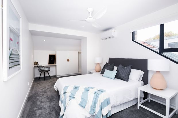 Bright contemporary bedroom with a large bed, dark upholstered headboard, white bedding, pale blue throw and cushions, matching bedside tables and lamps, grey carpet, built-in white wardrobes, a desk and chair, wall-mounted air conditioner, ceiling fan, and angled windows providing natural light.