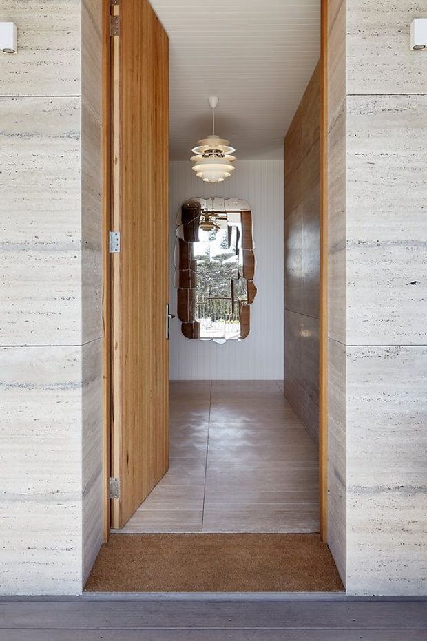 View through an open timber door into a narrow, refined interior passage or entry hall. The doorway is framed by horizontally jointed pale stone or concrete walls, with a warm timber door and timber wall panel on the right. Inside, large warm grey-brown floor tiles, vertically grooved pale wall panelling, a sculptural layered pendant light, and an organic-shaped decorative mirror create a calm mid-century-inspired composition. A dark timber threshold or deck is visible in the foreground.