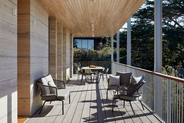 A spacious covered outdoor terrace or balcony with timber decking, a timber-lined soffit and wall, slender metal posts, and white metal balustrades. The terrace is furnished with woven lounge chairs, cushions, and a dining table and chairs, creating a relaxed entertaining area overlooking mature trees. Strong linear shadows and abundant daylight give the space a warm, airy contemporary feel.