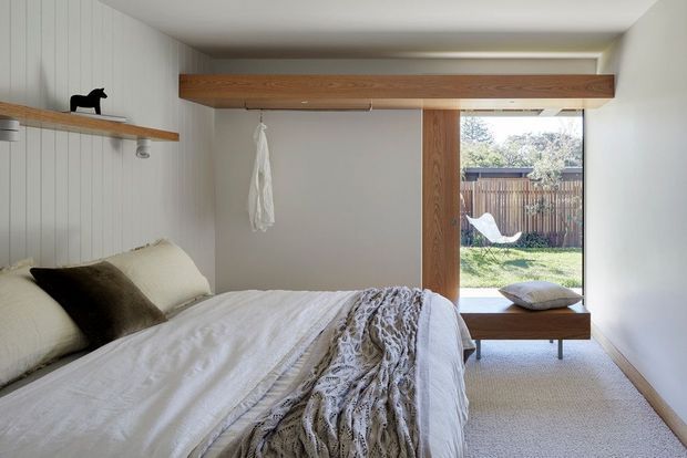 A compact, serene bedroom with a low-profile bed, white bedding, a textured knitted throw, and a dark accent cushion. White vertical wall panelling, pale carpet, and warm timber built-in shelving and overhead beam details create a simple, refined interior. A large glazed opening at the end of the room provides views to a fenced garden and a timber bench seat beneath the window.