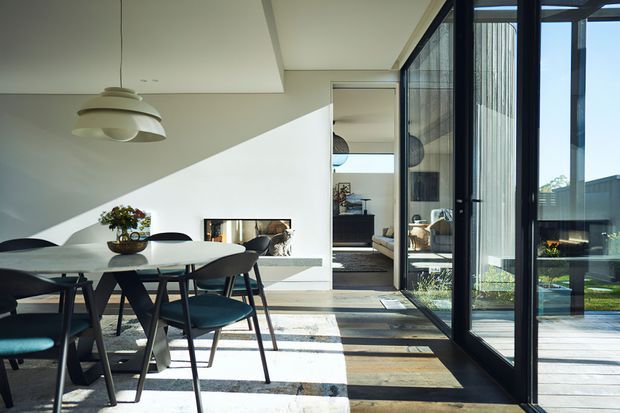 A bright contemporary dining area with a dark dining table and teal upholstered chairs beneath a sculptural pendant light. A low linear fireplace is set into a white wall beneath a sharply angled ceiling, while full-height glazed sliding doors open to a timber deck and landscaped exterior. The glazing and open interior connection create strong natural light and visual continuity with the outdoors.