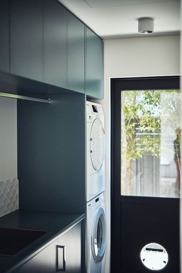 Compact, contemporary laundry room with dark blue-grey built-in cabinetry, a stacked white washer and dryer, dark benchtop, and a black-framed glazed exterior door. A small scalloped tile splashback is visible at the left edge, with recessed ceiling lighting and a pet door in the lower section of the door.
