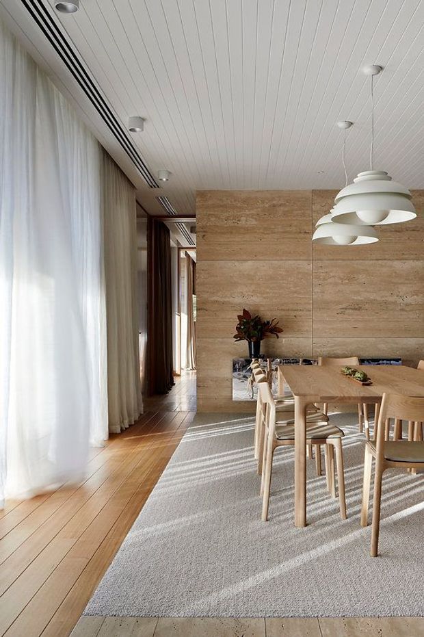 Bright, refined dining area with a long light timber dining table and matching chairs arranged on a large pale textured rug. Floor-to-ceiling sheer curtains line the glazed wall, while a warm beige travertine-like feature wall forms the backdrop. The white linear-panel ceiling includes recessed downlights and linear air-conditioning vents, complemented by sculptural white pendant lights. Timber flooring, soft daylight, and neutral natural materials create an airy contemporary interior.