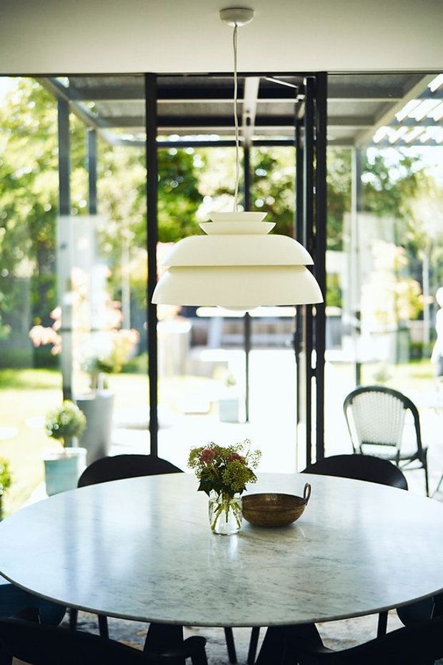 Bright dining area centred on a large round pale stone dining table with dark chairs, a sculptural white layered pendant light, and a small floral arrangement with a bowl. Full-height black-framed glazing opens the room visually to a leafy garden and outdoor seating. The space has a clean, light-filled contemporary character.