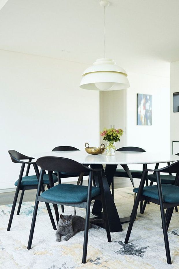 Bright, minimalist dining area with a round white dining table surrounded by black-framed chairs upholstered in deep teal. A large white layered pendant light hangs above the table, which is styled with a small vase of flowers and a decorative bowl. The room has white walls and ceiling, pale flooring, and a light patterned rug; a grey cat is seated beneath the table.