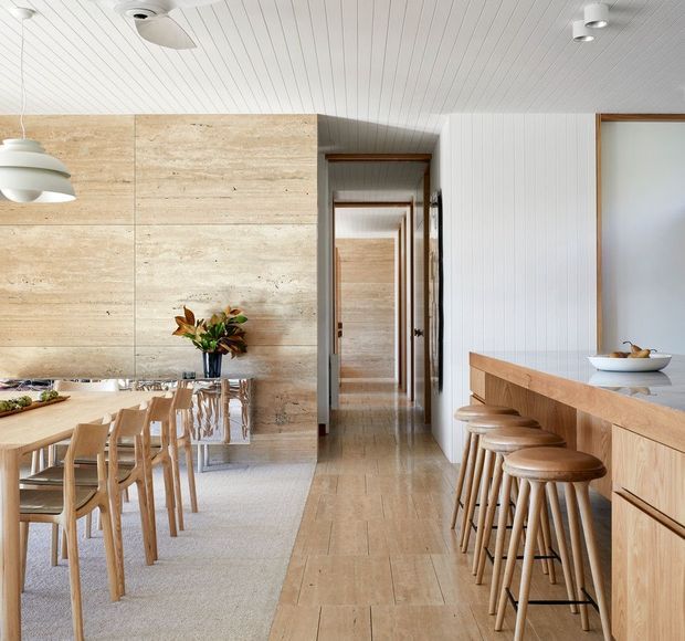 Bright open-plan dining and kitchen interior with a long timber dining table and chairs, a timber island with stools, pale natural stone wall cladding, light timber flooring and cabinetry, and a white vertically panelled ceiling and wall. A hallway extends through the centre of the home, with understated contemporary detailing and warm natural materials.