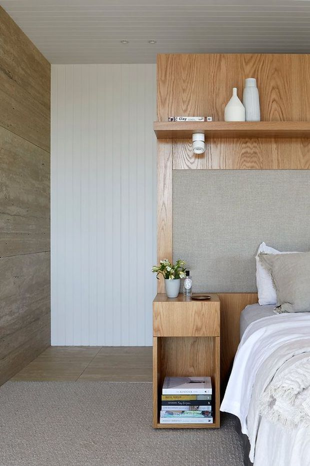A close-up view of a calm, contemporary bedroom interior. The image shows a partially visible bed with white layered bedding and a grey upholstered headboard, integrated into a warm timber feature wall with an open shelf and recessed spotlight. A timber bedside table with open book storage, a small plant and accessories sits beside the bed. The room combines vertical white wall panelling, a textured grey carpet, large-format grey floor tiles, and a raw concrete-look wall.