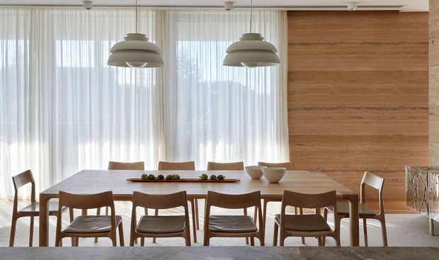 A bright, refined dining room with a long timber dining table surrounded by wooden chairs, sheer full-height curtains, sculptural layered pendant lights, and a warm horizontal timber feature wall. A simple low centerpiece and ceramic bowls sit on the table, creating a calm, natural, contemporary setting.