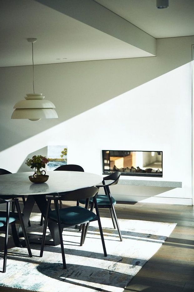 A bright, minimalist dining area with a round light stone or pale composite dining table surrounded by dark chairs. A sculptural white pendant light hangs above, while a sleek built-in linear fireplace is set into the white wall. Strong diagonal sunlight creates dramatic shadows across the walls and a large patterned rug, with dark timber flooring adding contrast.