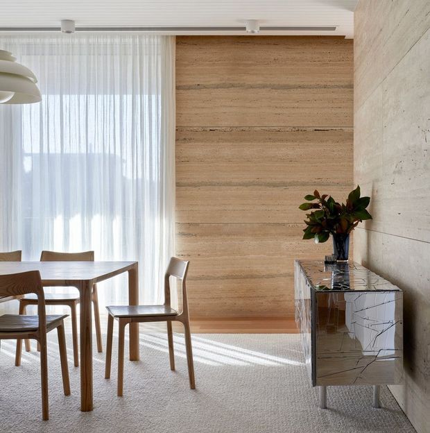 Bright, refined dining area with a timber dining table and chairs beside sheer floor-to-ceiling curtains. The room features a large horizontal light timber wall panel, a textured concrete wall, a ribbed white ceiling, a sculptural pendant light, and a mirrored geometric-front sideboard with a potted plant. Natural daylight creates a calm, contemporary atmosphere.