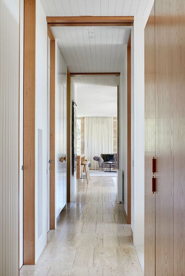 A bright, narrow interior hallway leading toward a furnished living area. The corridor features white vertical wall and ceiling panelling, pale stone or concrete-look tiled flooring, natural timber door frames and beams, and a large timber-clad door or wall panel on the right. A lounge area with seating and sheer curtains is visible at the far end.
