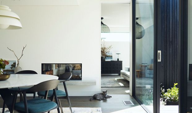 Bright contemporary open-plan dining and living area viewed from an entry or adjoining space. The foreground features a round white dining table with dark sculptural chairs, a large white pendant light, and a minimalist built-in fireplace or media niche. Sliding glass doors with dark frames open to a planted courtyard or outdoor area, while the background reveals a living room with a dark built-in cabinet and neutral furnishings. The palette is clean and restrained, combining white surfaces, pale flooring, dark timber or metal framing, and muted blue upholstery.