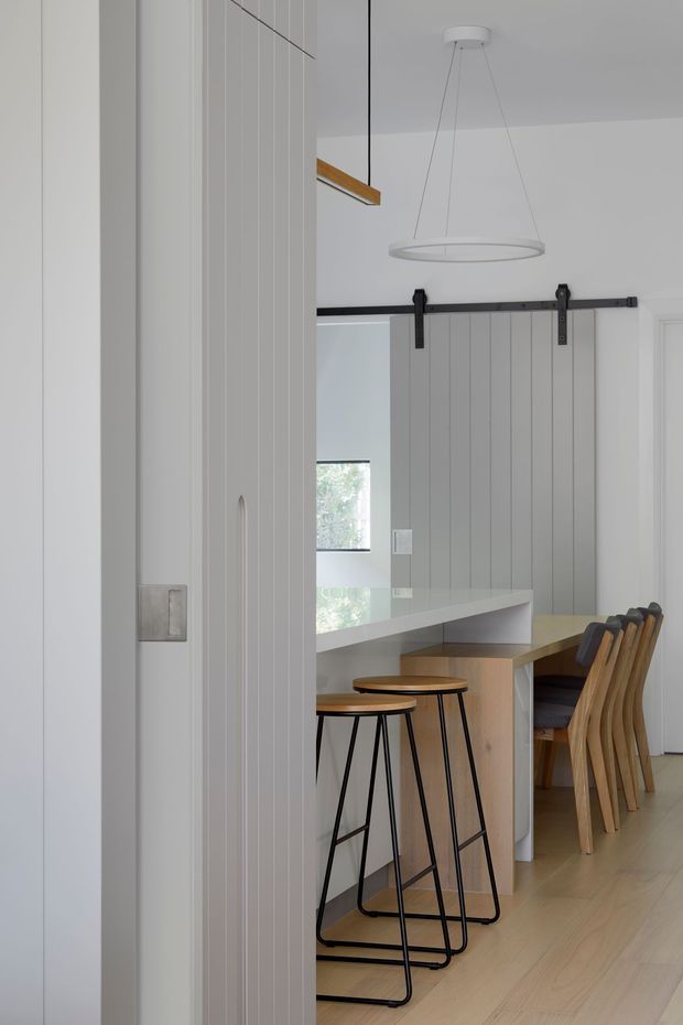 A bright, minimalist dining and kitchen area viewed through a partially open white panelled door. The room features a light timber island or dining counter, white cabinetry and vertical wall panelling, a black barn-door track with matching sliding panel, pale timber flooring, timber dining chairs, black metal stools, and a simple circular pendant light. The overall composition is clean and understated with warm timber accents and strong white surfaces.