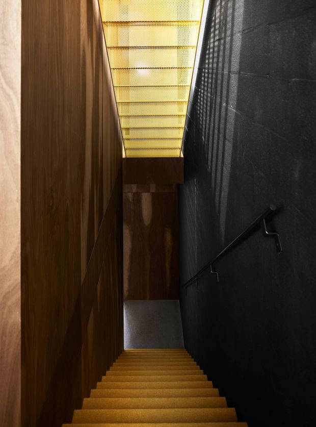 A narrow, dramatic interior stairway descends between dark timber wall panelling on the left and a textured black wall on the right with a simple black handrail. Warm golden stair treads lead toward a lower landing, while a translucent perforated ceiling or skylight brings in soft amber light.