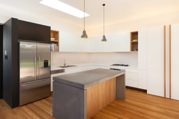 Bright contemporary kitchen with a large central island, white flat-panel cabinetry, dark integrated storage and stainless-steel refrigerator, warm timber flooring, a pale hexagonal-tile splashback, and two pendant lights. A skylight or rooflight adds natural illumination.