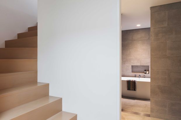 Interior view showing a light timber staircase beside a white partition wall, with a partially visible contemporary bathroom beyond featuring large grey-beige wall tiles, a built-in bathtub, recessed niche, and warm recessed lighting.
