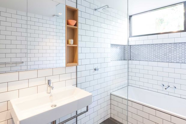 Bright contemporary bathroom featuring extensive white rectangular wall tiling, a freestanding white wall-mounted basin, frameless glass shower screen, and a built-in white tiled bathtub. A warm timber recessed shelf provides contrast, while a patterned hexagonal tile band adds a subtle feature detail. Natural light enters through a high horizontal window.