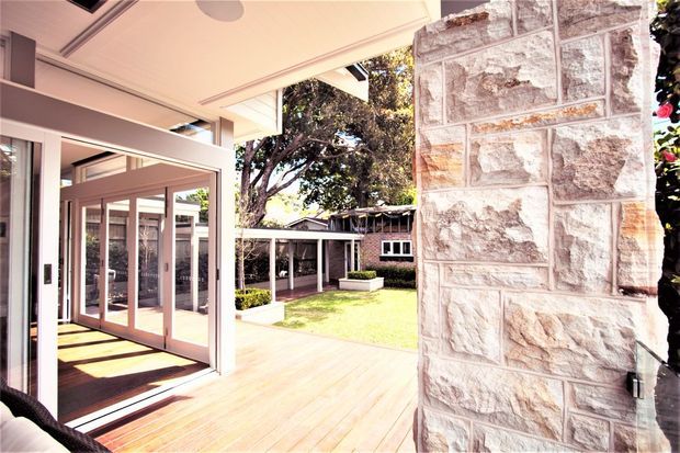 Bright covered outdoor terrace adjoining a glazed interior space, with a timber deck, large sliding glass doors, a substantial exposed stone wall, and views into a landscaped courtyard with a secondary low-slung structure. The setting combines contemporary glazing and canopy detailing with a textured masonry feature wall.