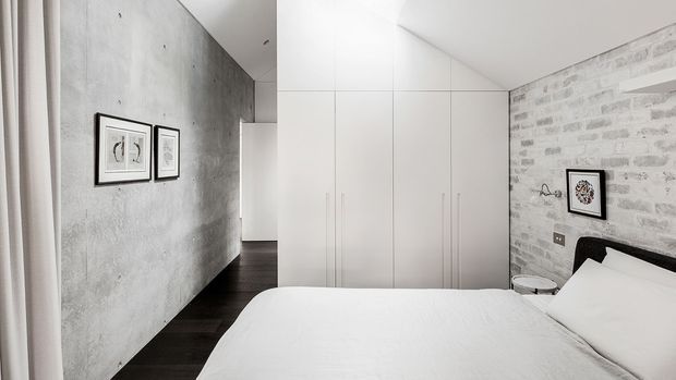 Contemporary minimalist bedroom with a low-profile bed, white bedding, full-height white built-in wardrobes, a dark timber floor, a raw concrete-look feature wall, and a whitewashed exposed brick wall. Small framed artworks and restrained furnishings add subtle detail.