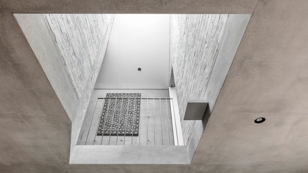 Upward view through a double-height interior void, showing exposed pale textured wall surfaces, a minimalist white ceiling, a recessed downlight, and a simple metal balustrade around the upper-level opening. The composition emphasizes the vertical connection between levels and the contemporary architectural detailing.