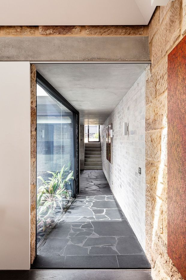A narrow, linear interior passageway with a dark irregular stone floor, exposed concrete ceiling, pale painted masonry wall, and warm rough stone wall framing. Full-height black-framed glazing runs along one side, revealing planted space and bringing in natural light. Steps lead upward toward a bright opening at the far end, creating a strong axis through the corridor.