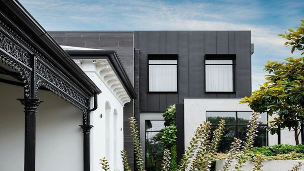 Exterior architectural view showing a contemporary dark-panelled two-storey building adjoining a more ornate heritage-style facade. The modern portion has broad charcoal cladding, simple rectangular windows with projecting dark frames, and white rendered lower walls, while the adjoining structure features elaborate black decorative metalwork, columns, moulded detailing, and a white eaves soffit. Established landscaping partially frames the buildings.