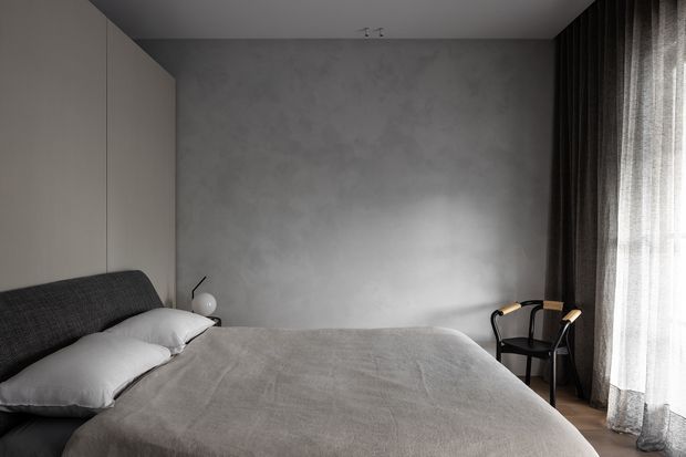 A serene, minimalist bedroom with a broad upholstered bed dressed in grey bedding, a textured grey plaster feature wall, pale built-in wardrobes, and a black accent chair beside floor-to-ceiling sheer and dark curtains. The room has a restrained monochromatic palette and soft natural light.