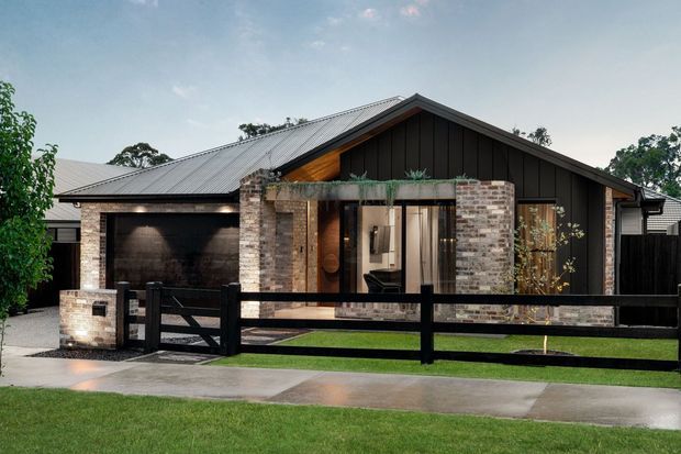 Street-facing exterior view of a single-storey contemporary home with a prominent dark gabled roof and vertical dark cladding, mixed brick masonry walls, a covered entry, and an integrated double garage. A black timber-style fence, paved path, small lawn, and restrained landscaping frame the facade.