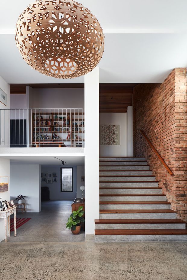 Interior view of a multi-level home centered on a stairway with timber treads, a red brick feature wall, and a timber-lined ceiling. A large decorative spherical pendant hangs above a polished concrete floor, with an open mezzanine, metal balustrade, shelving, and adjoining living area visible.